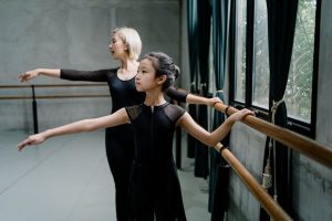 Home Ethnic girl in uniform with professional ballerina working out near barres while looking away in hall
