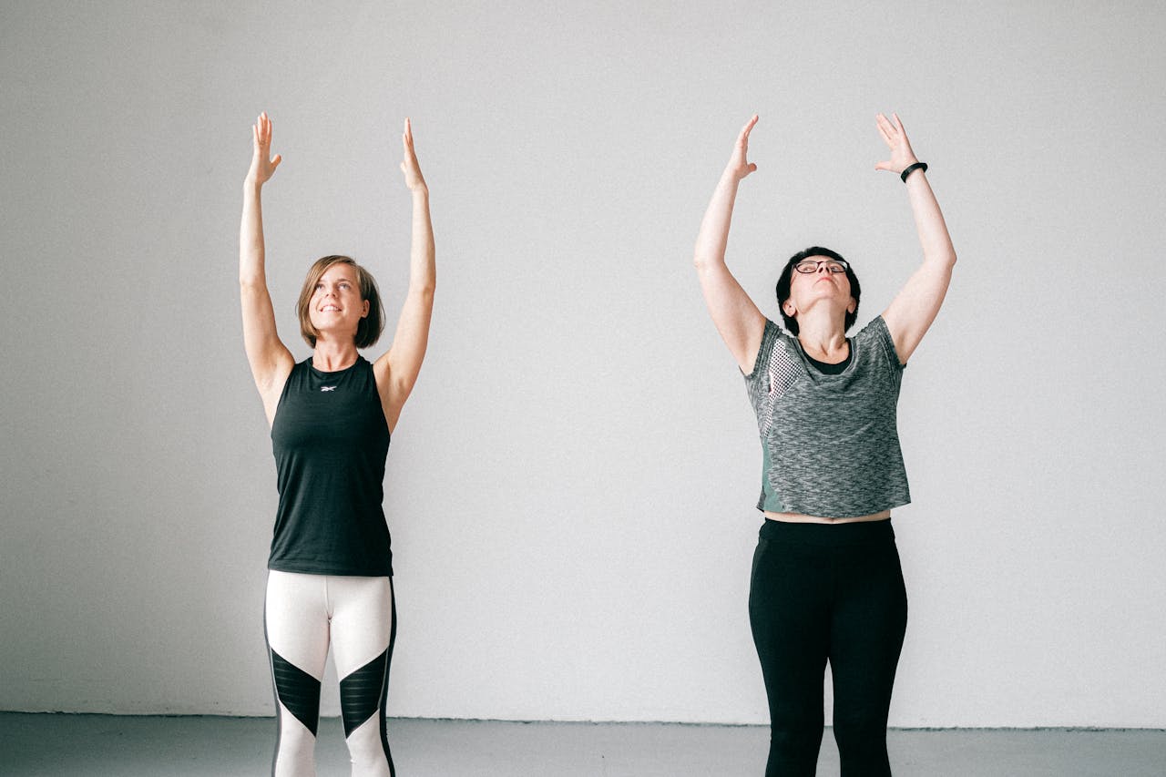 Home Two women engaged in a yoga session indoors, standing with arms raised, promoting fitness and wellness.